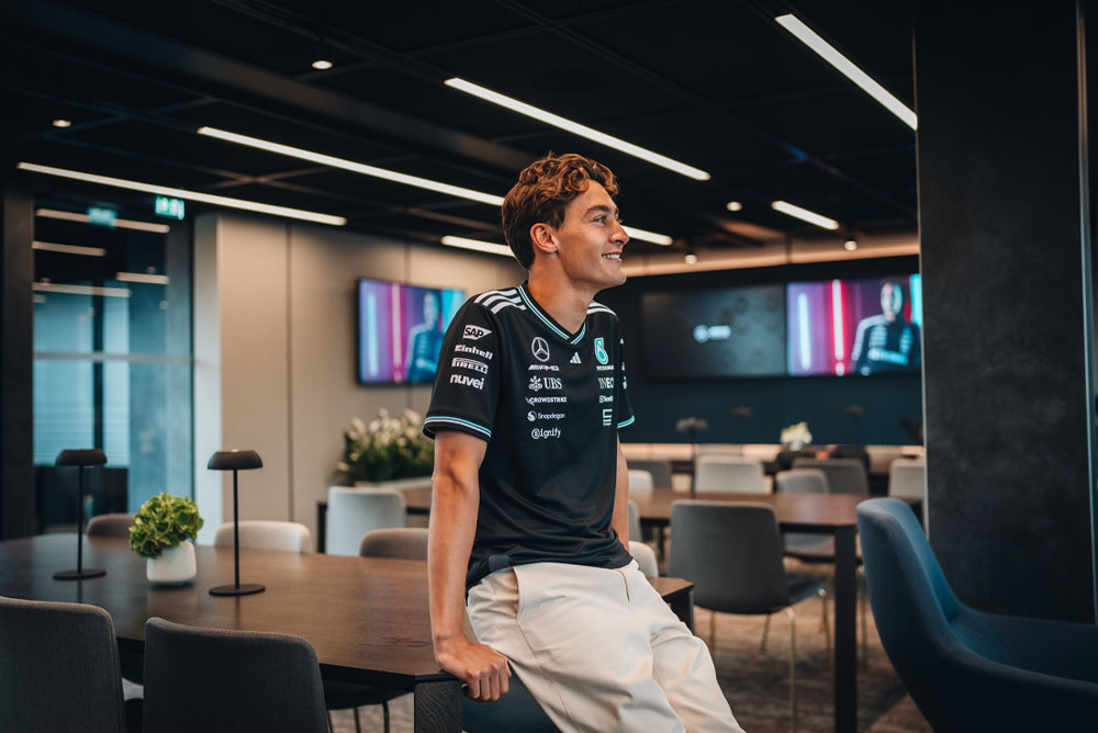 Джордж Ръсел, пилот на отбора Mercedes-AMG PETRONAS Formula One Team, седи небрежно върху маса в модерна и стилна зона за гости или офис на отбора. Той се усмихва и носи официалната отборна риза с видими логота на спонсори като Einhell, IWC и CrowdStrike. Екраните на заден план показват неговия образ.