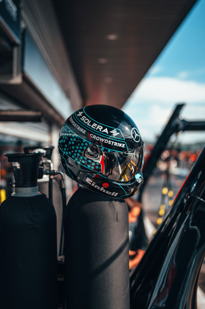 Каска на отбора Mercedes-AMG PETRONAS Formula One Team с ясно видими логота на спонсори, включително Einhell, CrowdStrike и Solera, е поставена върху газова бутилка в питлейна. Отражението върху визьора показва обстановката в гаража.