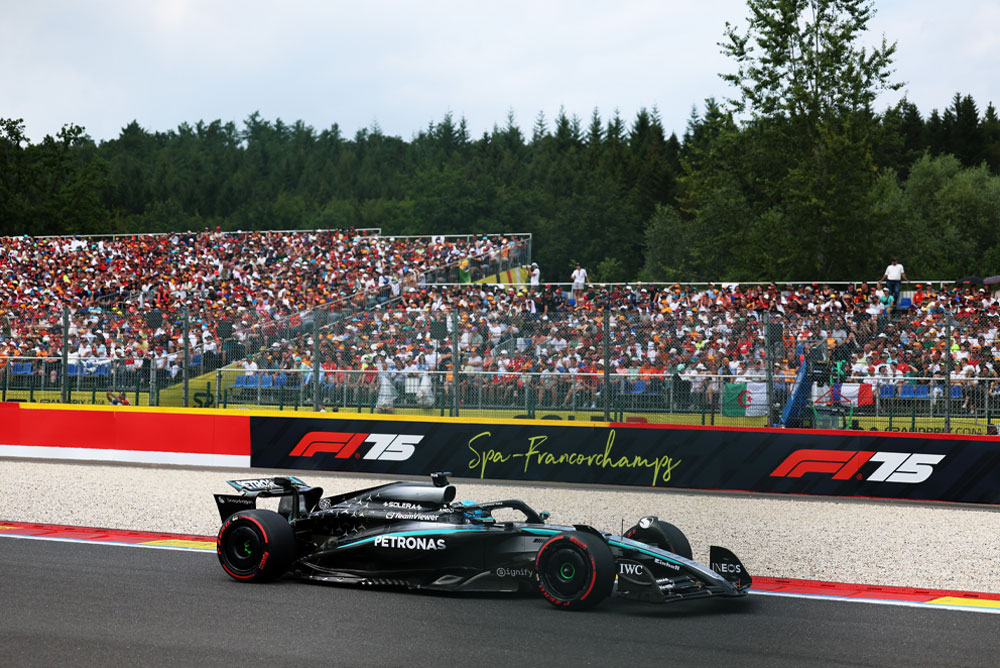 Автомобил на Mercedes-AMG PETRONAS Formula One Team се състезава на пистата Спа-Франкоршамп. На заден план трибуните са пълни с фенове, размахващи международни знамена. Логото на Спа-Франкоршамп е показано на бариерата.