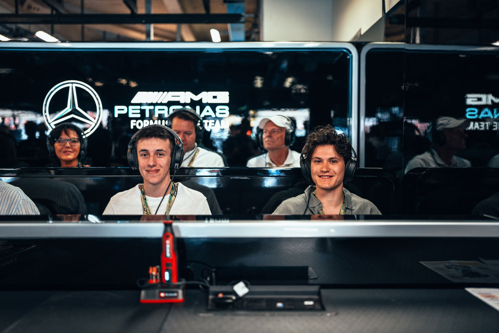 Двама мъже седят в гаража на Mercedes-AMG PETRONAS Formula One Team, носят слушалки и се наслаждават на преживяването зад кулисите. На преден план се вижда червено устройство Einhell.