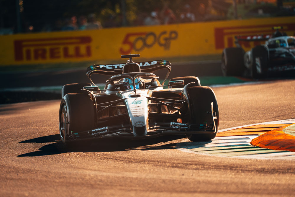 Автомобил на Mercedes-AMG PETRONAS Formula One Team се състезава по слънчева писта по време на Гран При. Колата завива с висока скорост, а на заден план се вижда друг автомобил.