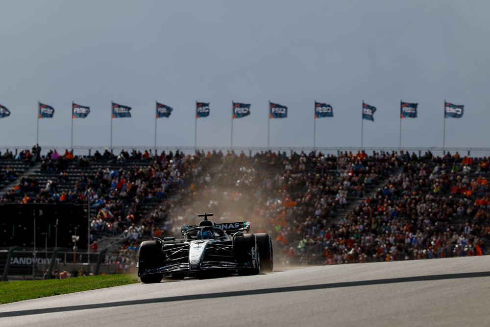 Автомобил на Mercedes-AMG PETRONAS Formula One Team преминава покрай пълни трибуни по време на състезание. Фенове размахват знамена на заден план, създавайки оживена атмосфера.