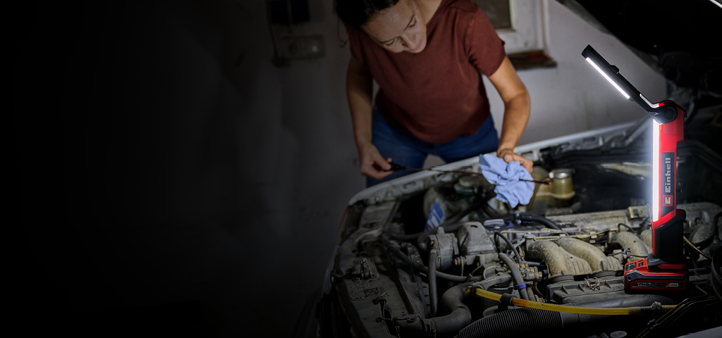 The image shows a person working on a car engine in a dimly lit garage. A bright red and black Einhell work light illuminates the engine area, providing clear visibility while the person uses a cloth and tools.